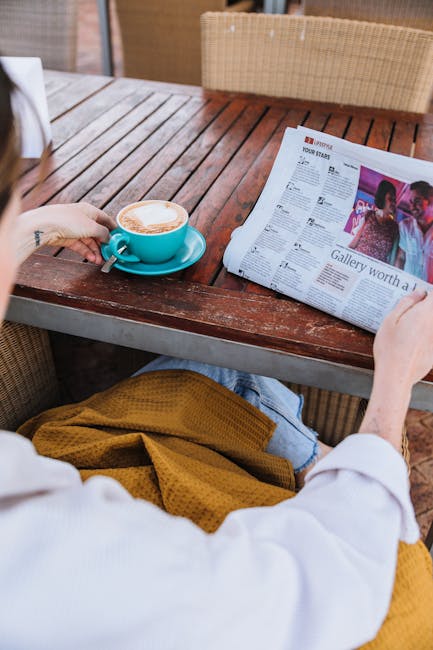 A person enjoys coffee while reading a newspaper at a wooden table.